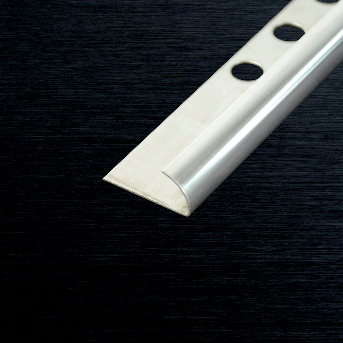 A close-up of a silver metal edge corner profile with three pre-drilled mounting holes, shown diagonally on a dark textured background.