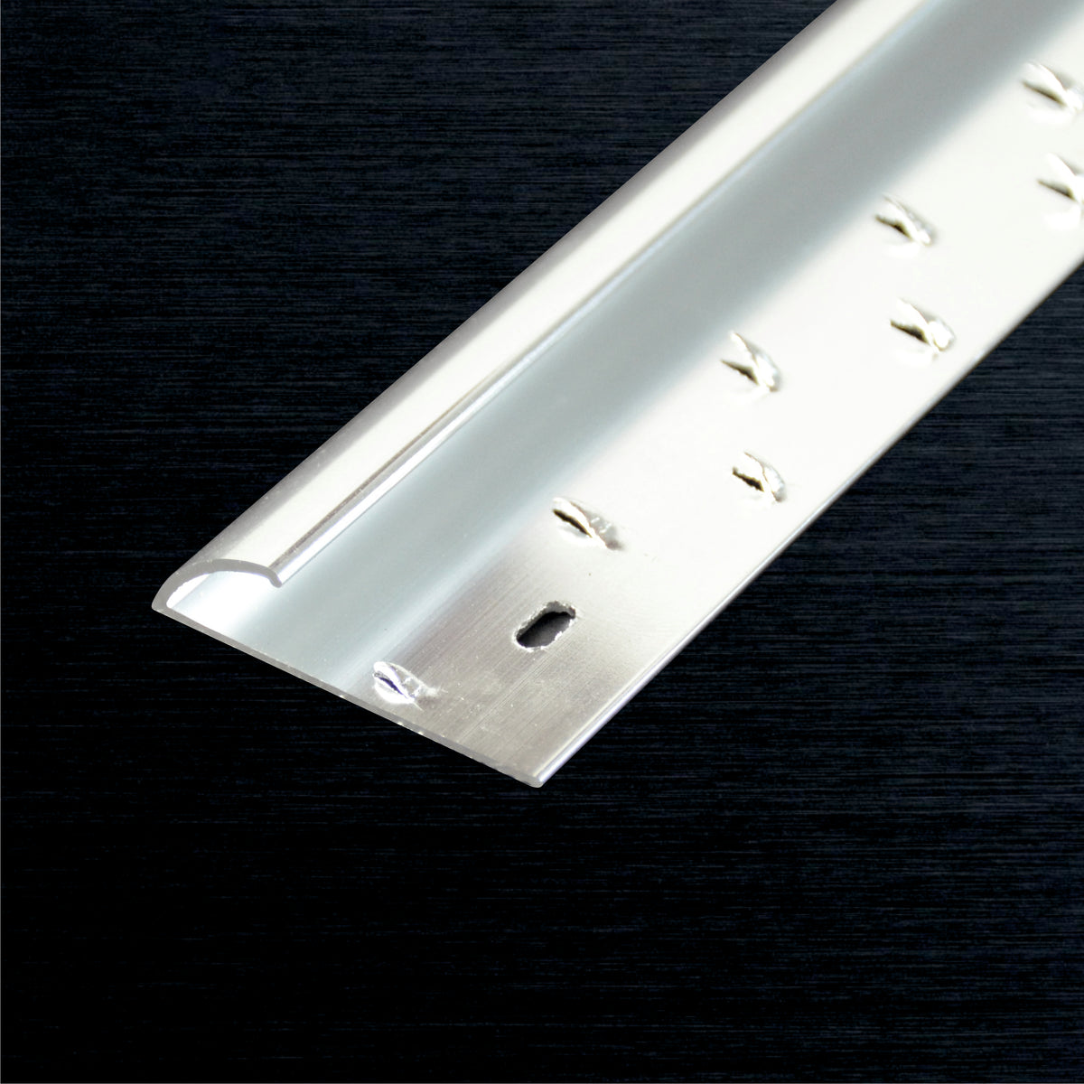 A close-up of a metallic silver aluminum carpet edge profile with pre-drilled screw holes, shown diagonally on a dark textured background.