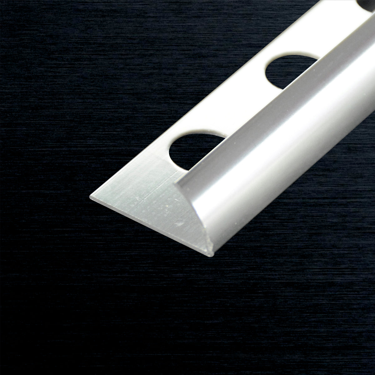 A close-up of a silver metal aluminum tile edge corner strip with two circular cutouts, shown diagonally on a dark textured background.