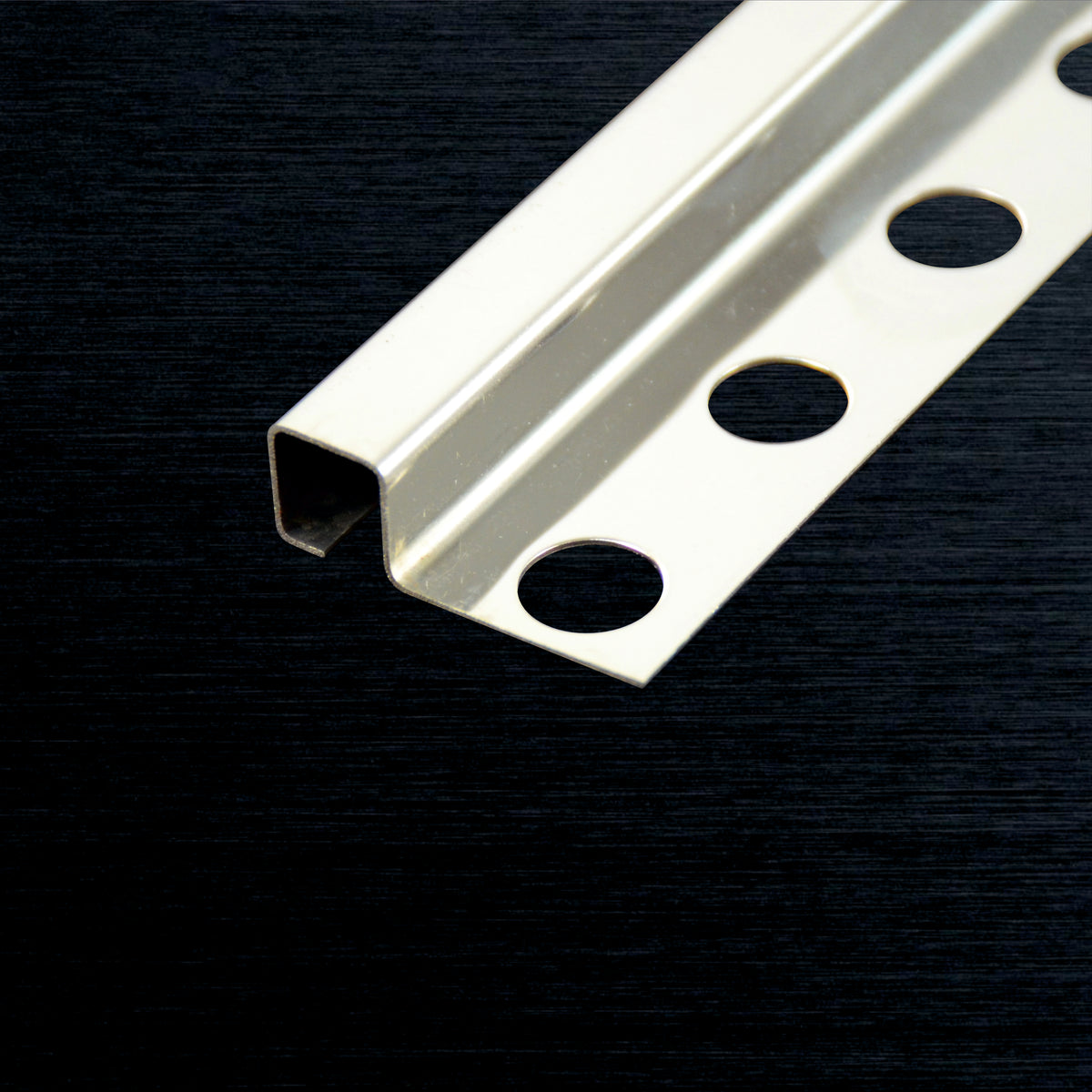 A close-up of a silver metal Tile Corner SS Square Profile with four circular mounting holes, shown diagonally on a dark textured background.