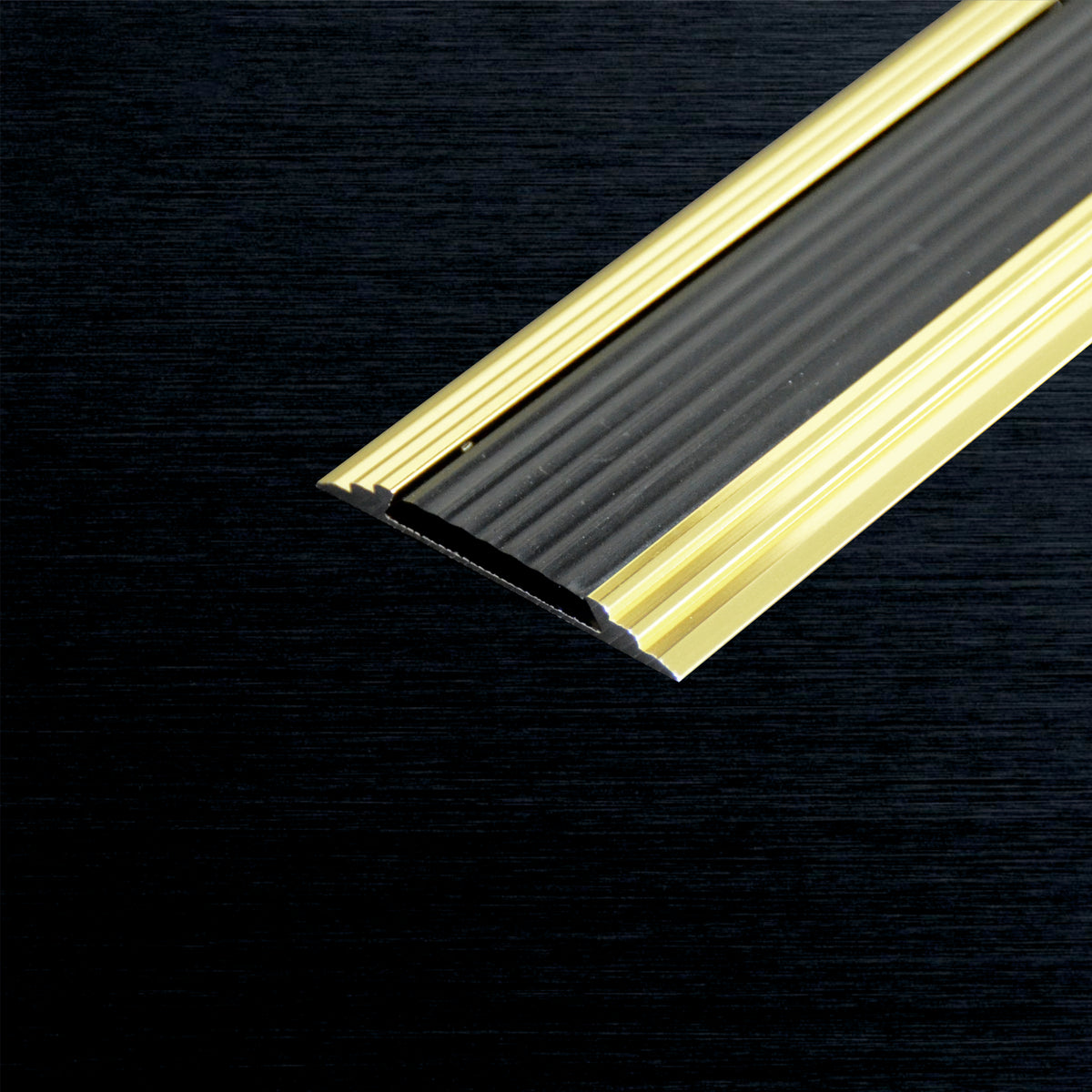 A close-up of a cover nosing profile strip featuring a brushed gold metallic outer edge and a ribbed black textured inner surface, shown on a dark wood background.