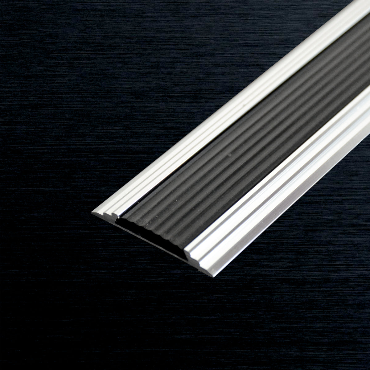 A close-up of a metal cover nosing profile piece with a ribbed black center and silver outer edges, shown diagonally against a dark textured background.