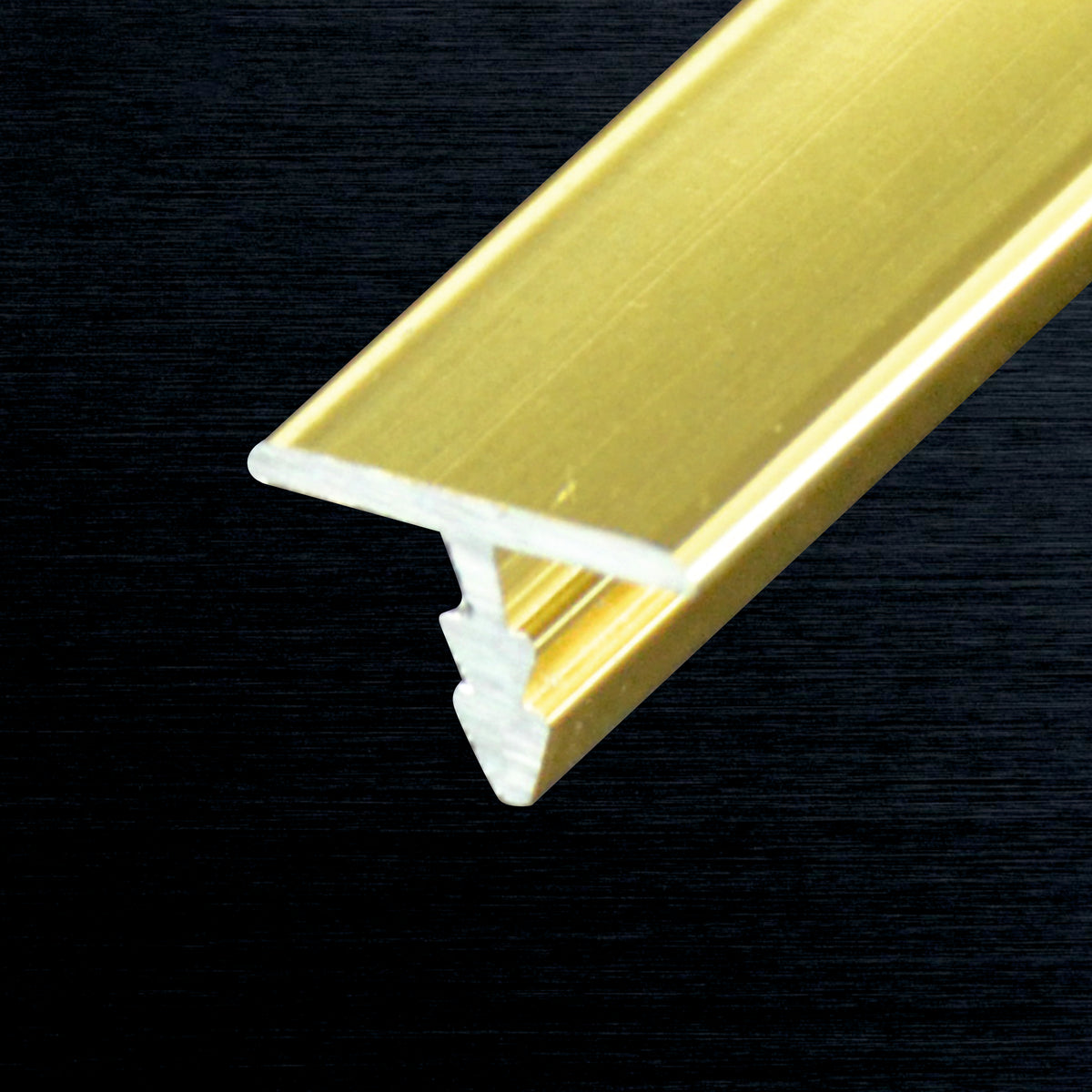 A close-up of a gold-colored metal T-profile with a white inner backing, shown diagonally against a dark textured background.