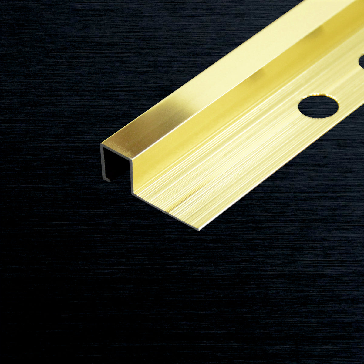 A close-up of a brushed gold metal square tile profile with a pre-drilled mounting hole, set against a dark textured background.