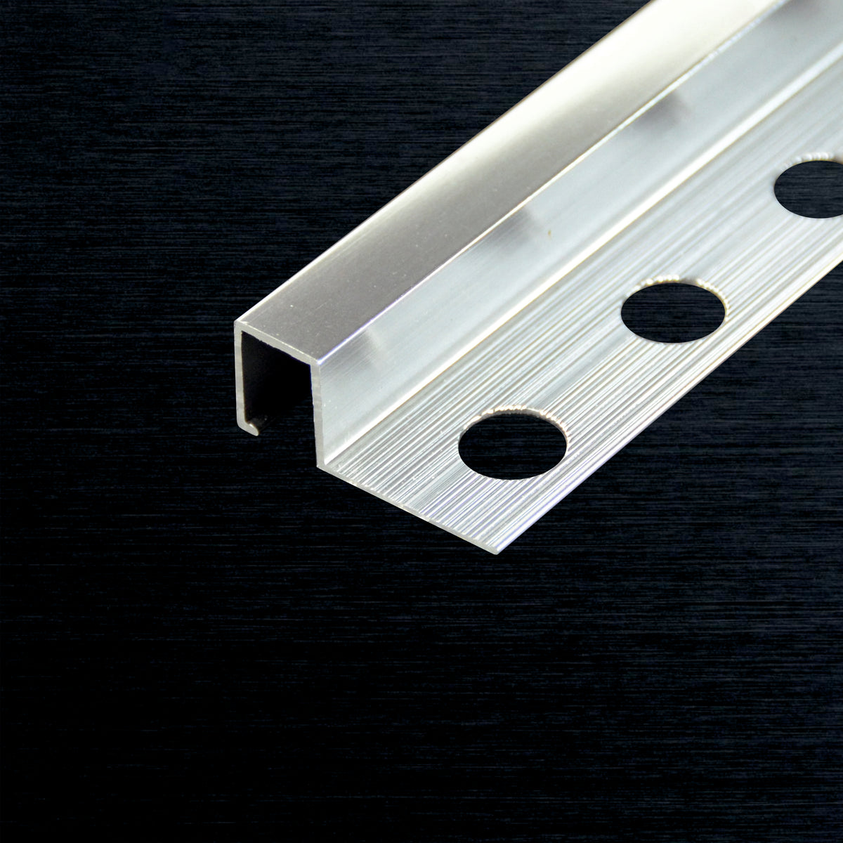 A close-up of a silver aluminum square tile profile with three pre-drilled circular holes, shown on a dark textured background.