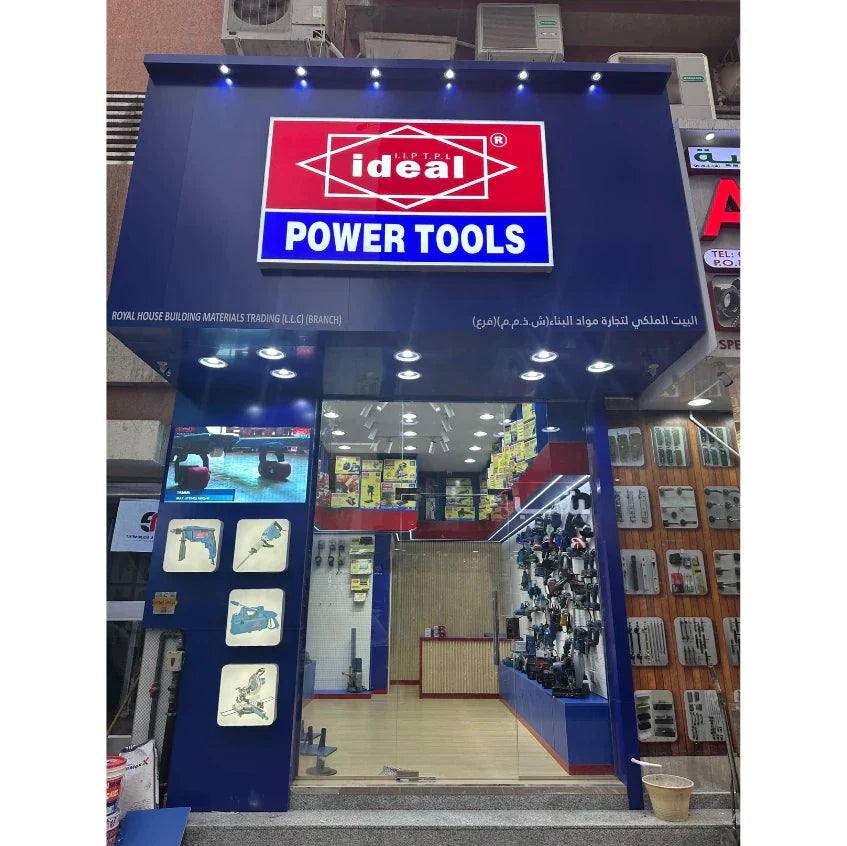A close-up, eye-level shot of the brightly lit "Ideal Power Tools" storefront at its grand opening, featuring a "GRAND OPENING" banner, red velvet ropes, and an inviting interior showcasing organized tool shelves under a warm, golden light.