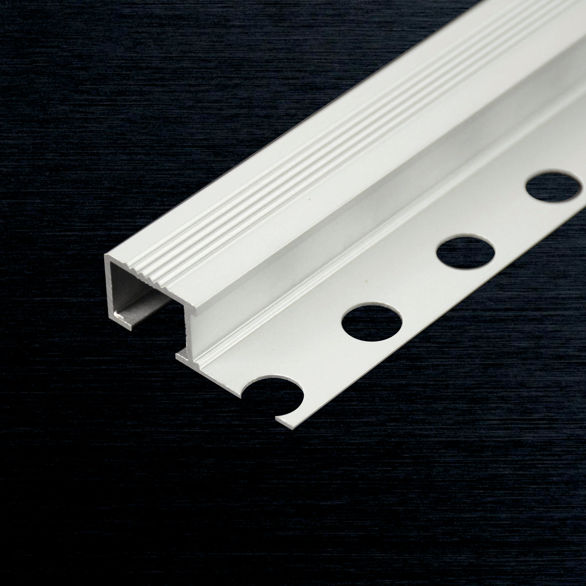 A white aluminum  stair grip profile with a ribbed top surface and three circular mounting holes, shown diagonally on a dark textured background.