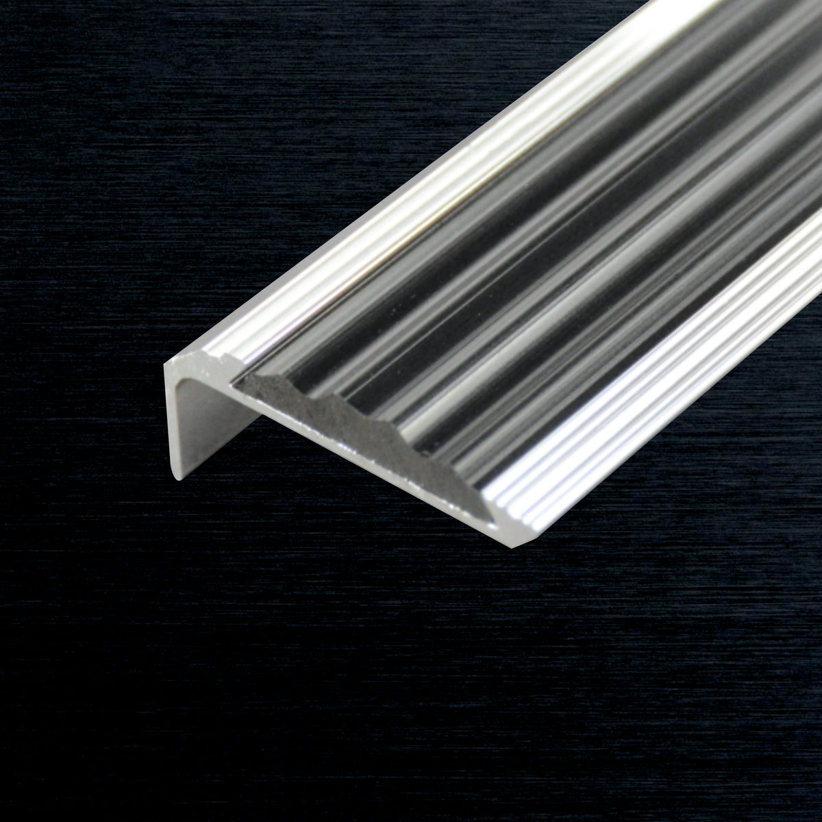 A close-up of a silver aluminum stair nosing profile piece with a ribbed interior, shown diagonally against a dark textured background.