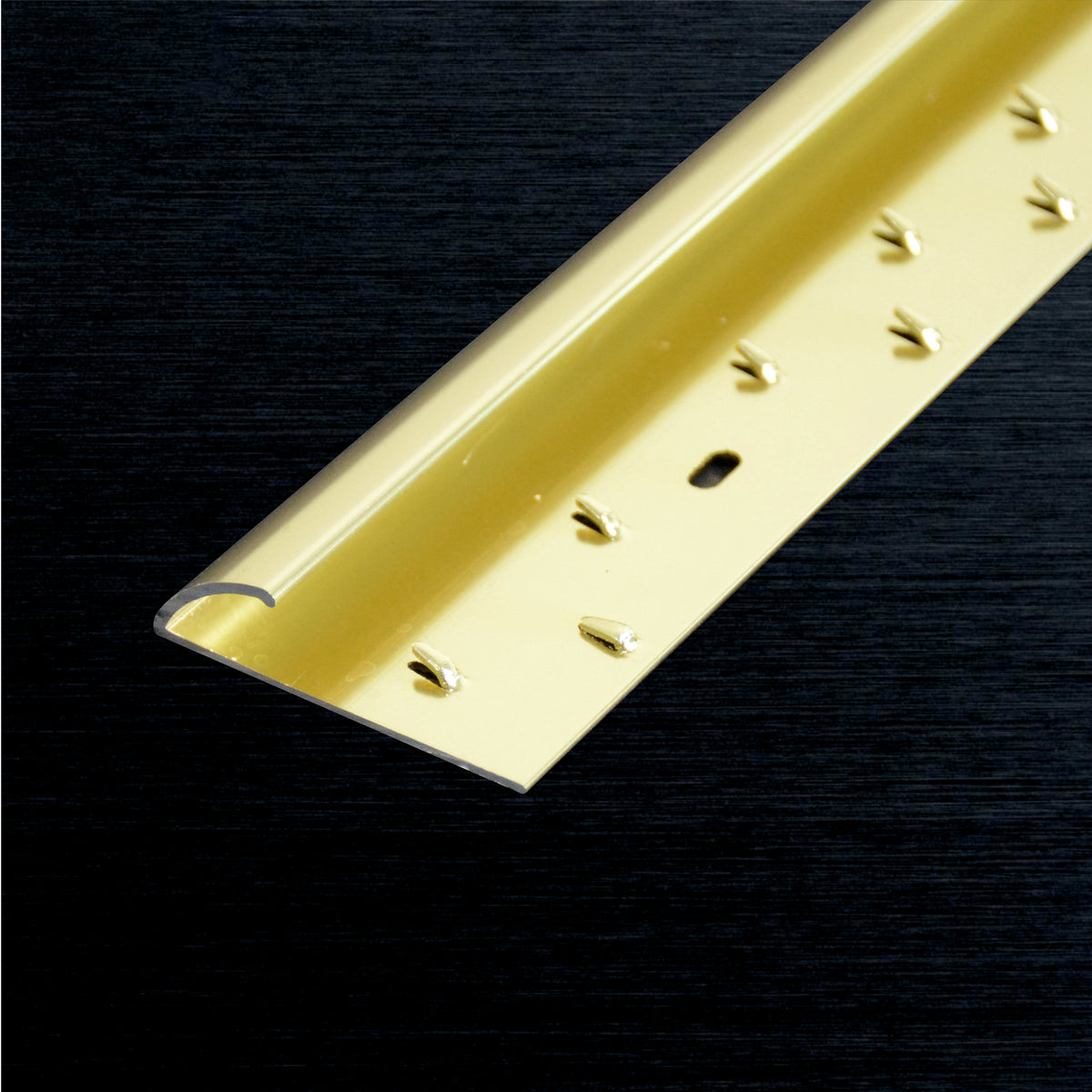 A close-up of a metallic gold aluminum carpet edge profile piece with pre-drilled holes, shown diagonally on a dark textured background.