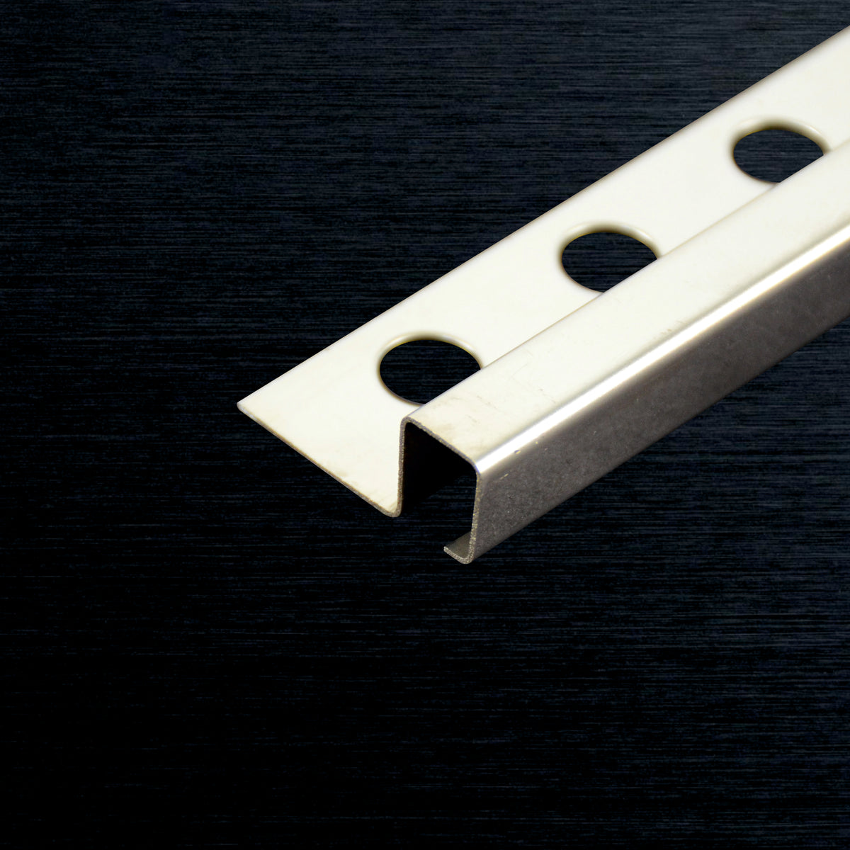 A close-up of a tile corner profile IDP-014-10HB with three circular mounting holes, shown on a dark textured background.
