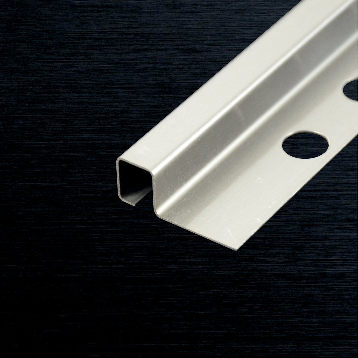 A close-up of a tile corner profile with a pre-drilled mounting hole, shown diagonally on a dark textured background.