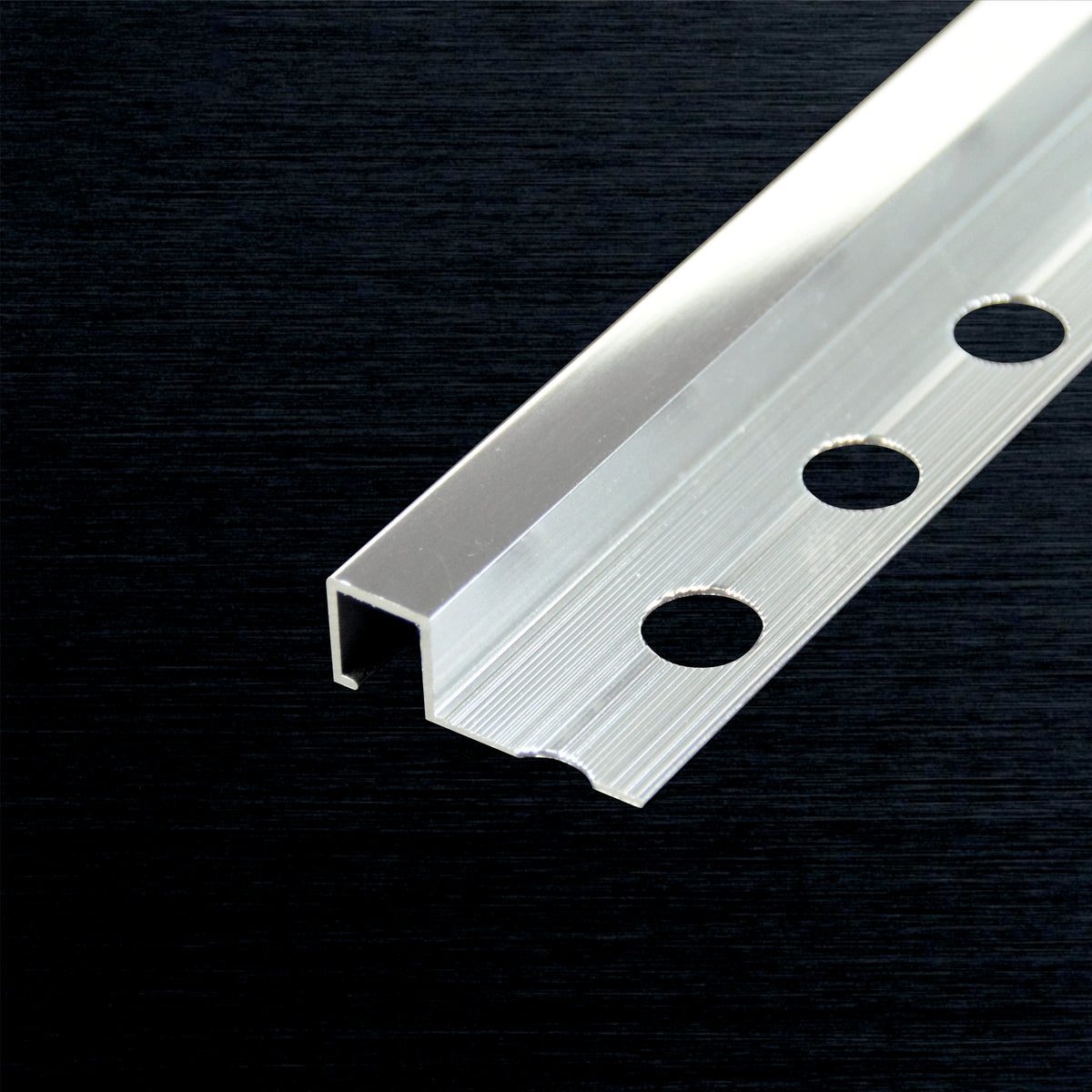A close-up of a silver aluminum square tile profile with three circular mounting holes, shown diagonally on a dark textured background.