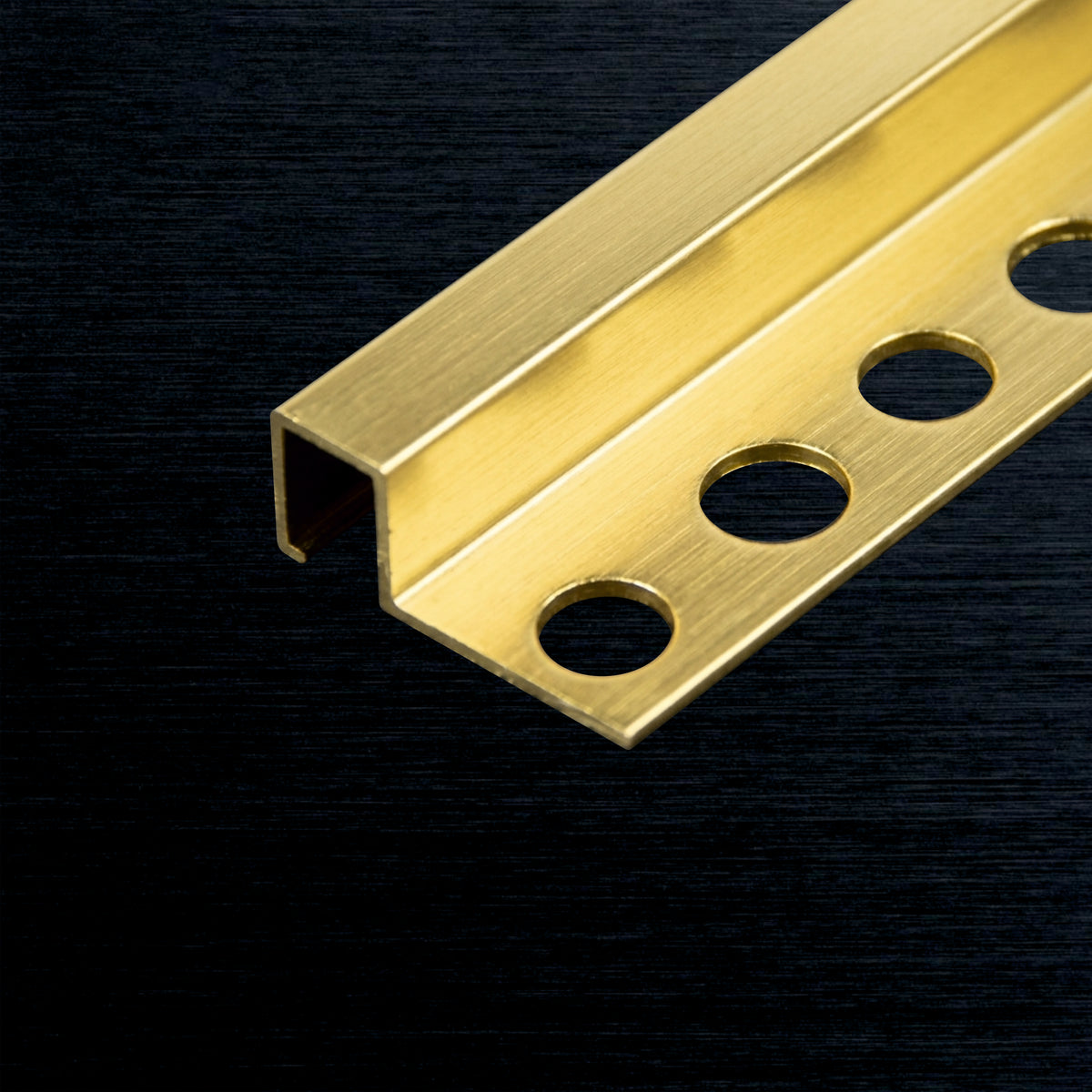 A close-up of a brushed brass or gold-colored square tile profile strip with four circular mounting holes, shown diagonally on a dark textured background.