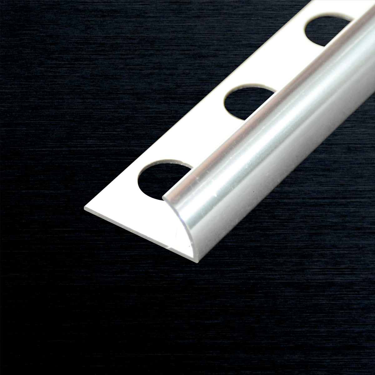 A close-up of a silver metal  tile edge corner profile piece with three circular cutouts, shown diagonally on a dark textured background.